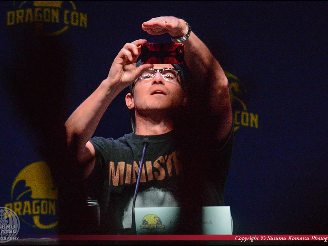 In this picture we can observe a man wearing a T shirt and holding a mobile phone in his hand. He...