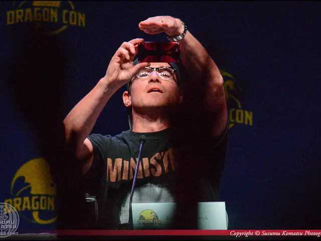 In this picture we can observe a man wearing a T shirt and holding a mobile phone in his hand. He...