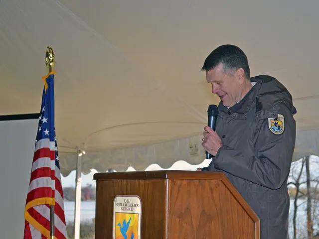 In this image we can a person is standing near a podium. A man is holding a microphone and speaking...