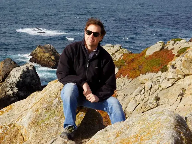 In this image I can see a man wearing a jacket, sitting on a rock and giving pose for the picture....